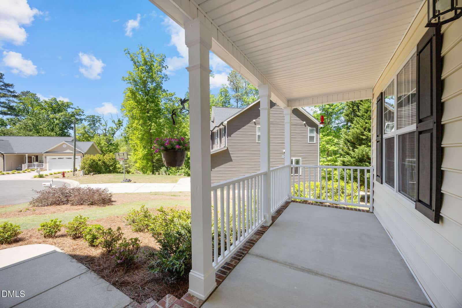 A view from a covered front porch with white railings, looking out onto a quiet suburban street with houses, trees, and a hanging flower basket on a sunny day.