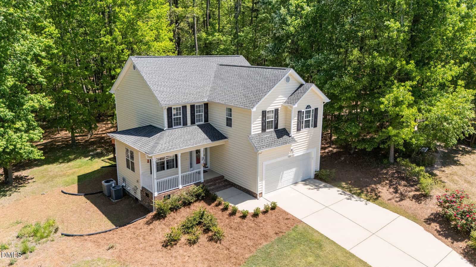 A two-story light yellow house with a gray roof, white trim, front porch, and attached two-car garage. The house is surrounded by trees and has a driveway and landscaped yard.