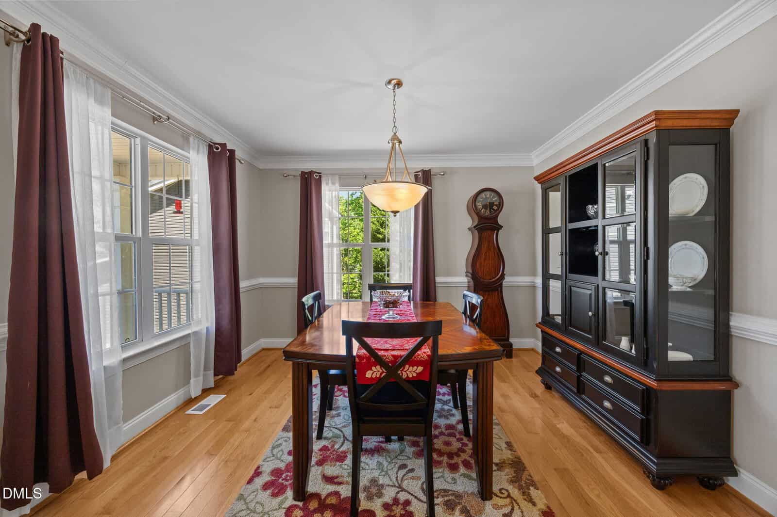 A bright dining room features a wooden table with six chairs, a floral rug, a china cabinet, a grandfather clock, and large windows with white and burgundy curtains.
