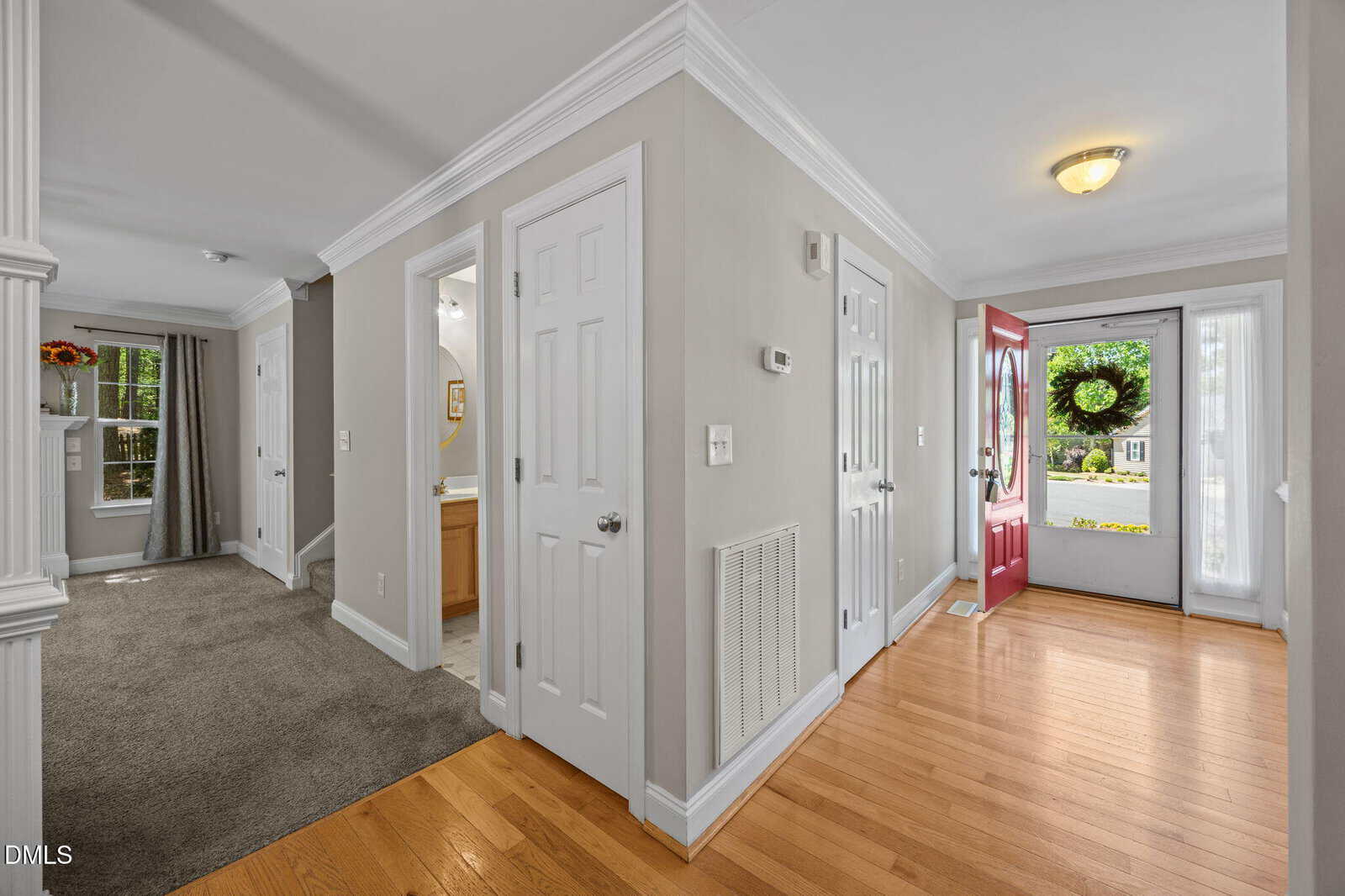 Bright entryway with hardwood floors, a red front door, white trim, and neutral walls. Doors lead to carpeted rooms and a bathroom. Windows bring in natural light; greenery is visible outside.
