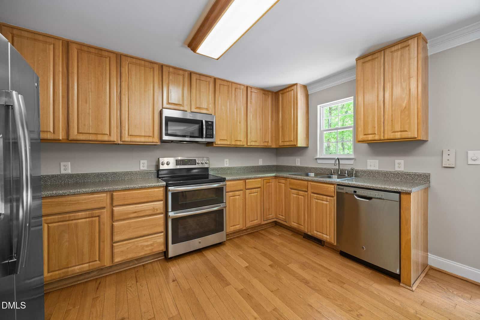 Modern kitchen with light wood cabinets, stainless steel appliances including a double oven, microwave, and dishwasher, granite countertops, a window above the sink, and hardwood floors.