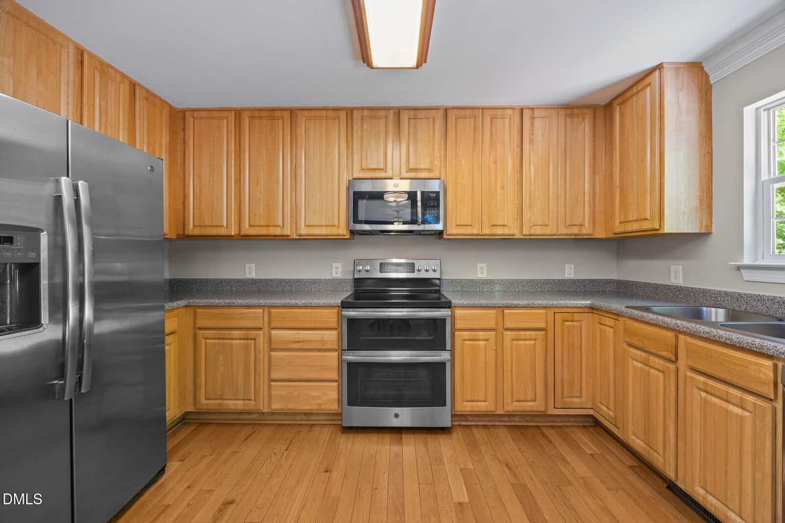 Modern kitchen with light wood cabinets, stainless steel appliances including a fridge, oven, and microwave, gray countertops, hardwood floors, and a window allowing natural light.