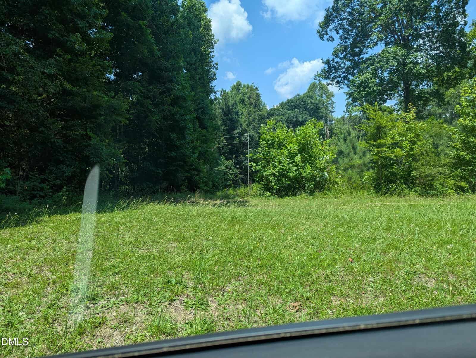 A grassy clearing bordered by dense green trees under a bright blue sky with scattered clouds, just outside 275 Perrytown, Warrenton. Part of a car windshield and dashboard are visible at the bottom edge of the photo.