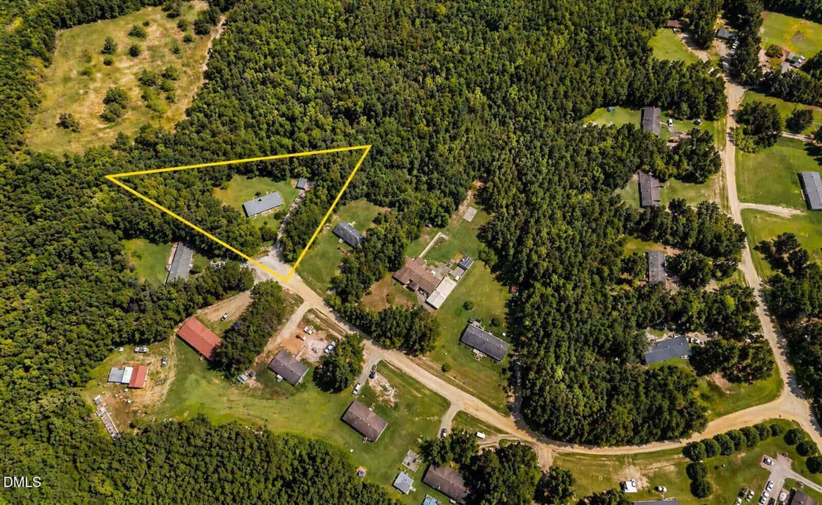 Aerial view of a rural neighborhood with several homes surrounded by dense trees. One triangular wooded lot is outlined in yellow, highlighting a specific property with a driveway and a house. Dirt roads connect the houses.