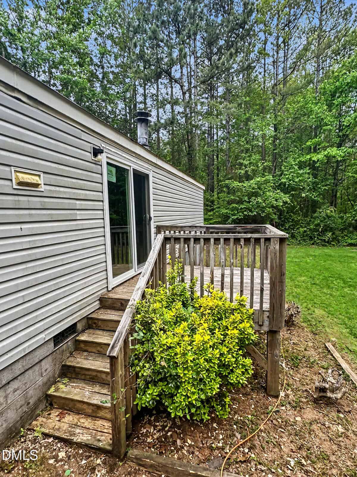 A small wooden deck with steps leads to a sliding glass door on a gray house. A green shrub grows beside the steps, and tall trees create a dense, forested background.