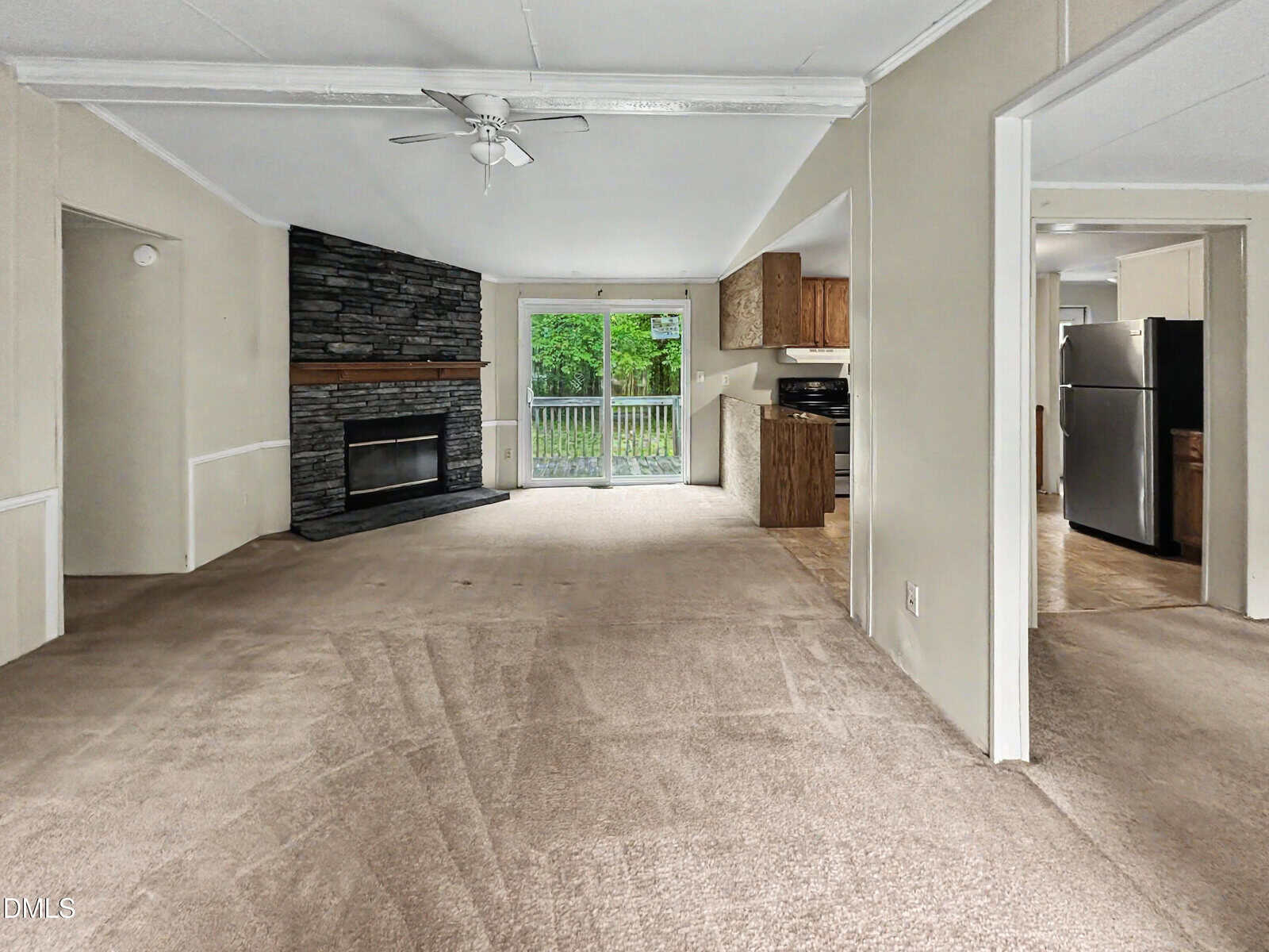 Living room with beige carpet, stone fireplace, ceiling fan, sliding glass doors to a patio, and an open view into the kitchen with wood cabinets and stainless steel appliances.