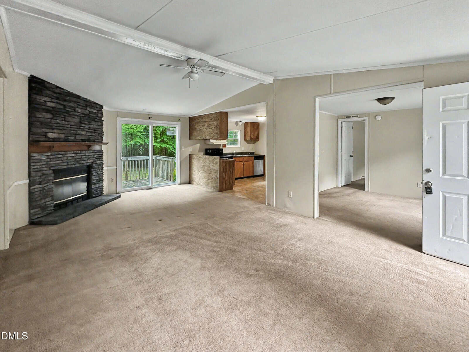 Open living room with stone fireplace, beige carpet, sliding glass doors to a deck, and a view into the kitchen with wood cabinets. A bedroom is visible through an open doorway on the right.
