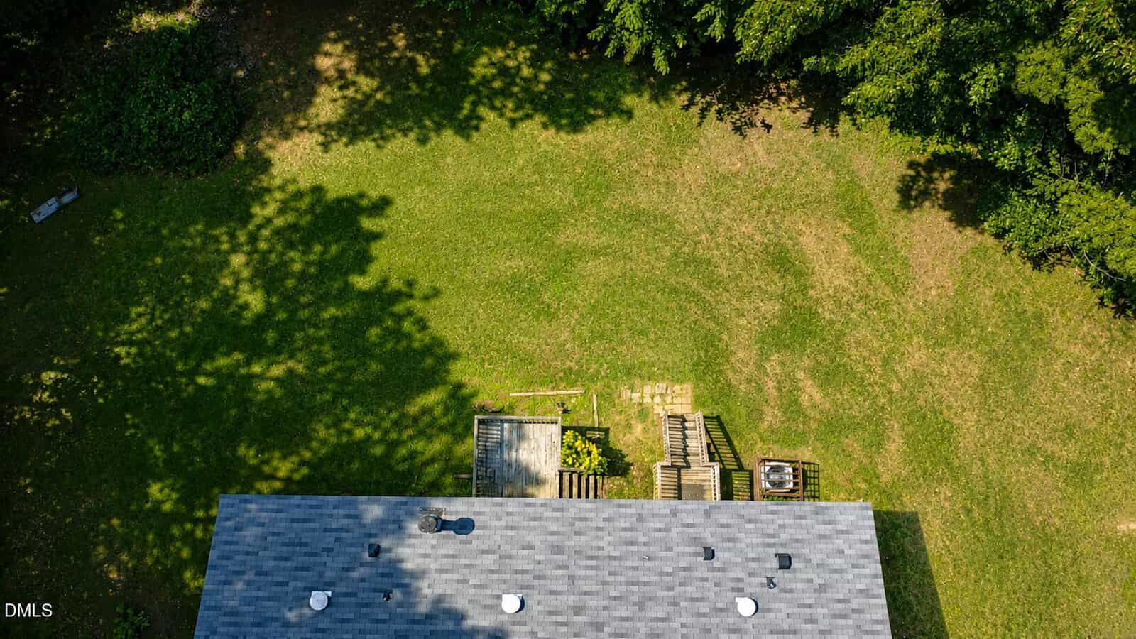 Aerial view of a grassy backyard bordered by trees, with the roof of a house and a wooden deck with stairs leading down to the lawn. Patches of sunlight and shadow cover the yard.