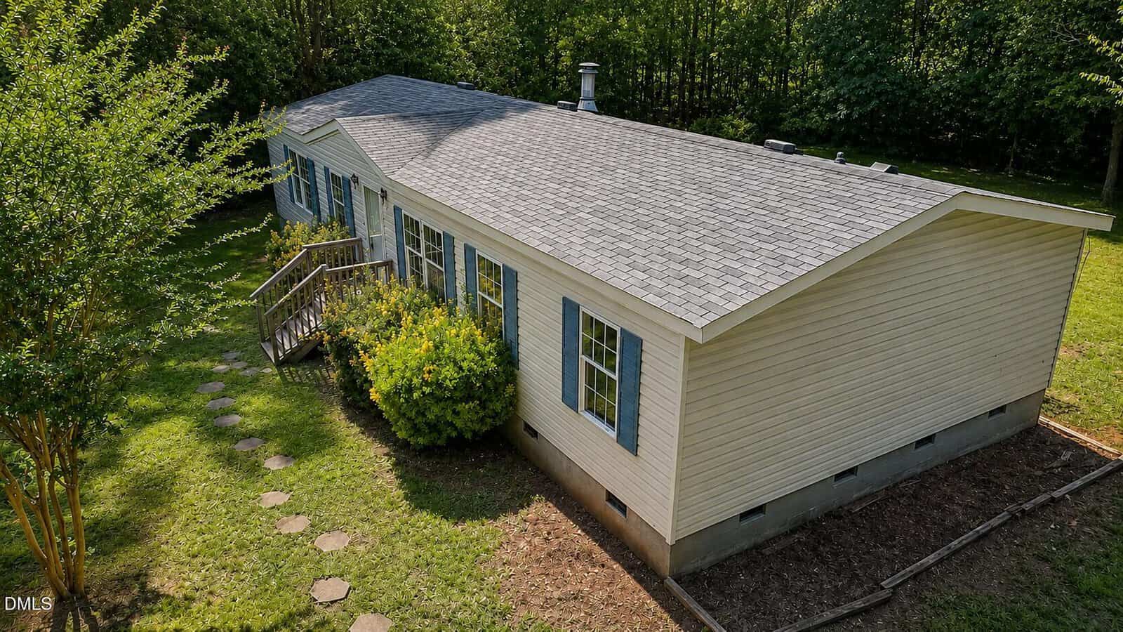 A light yellow single-story manufactured home with blue shutters and a gray shingle roof, surrounded by green grass, bushes, trees, and a stone walkway leading to the front porch.