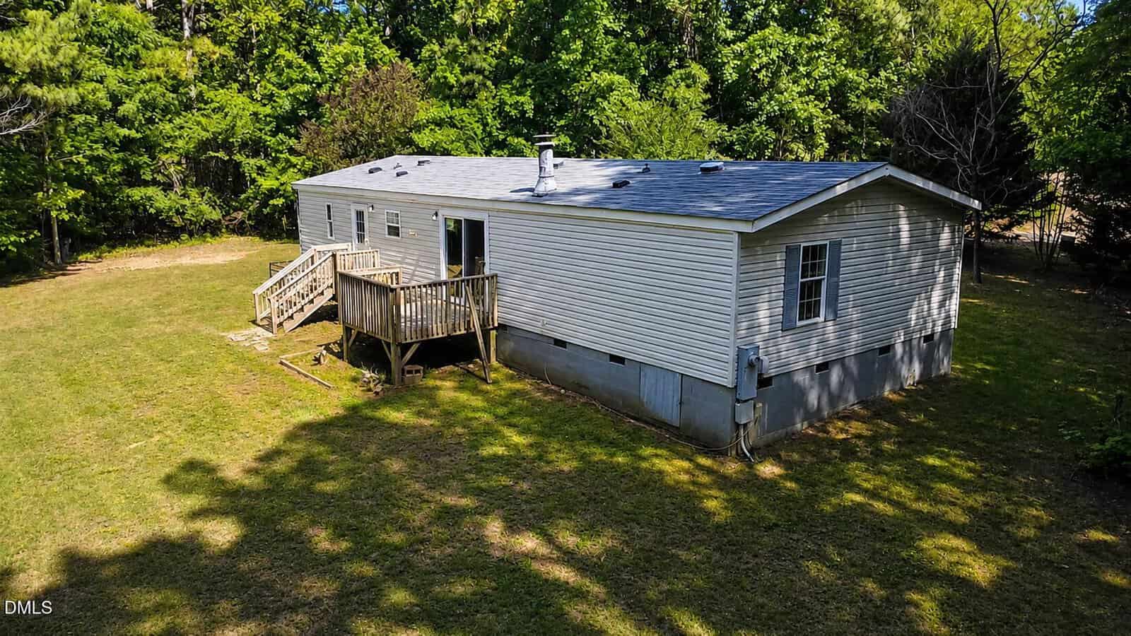A single-story manufactured home with light-colored siding, a small wooden deck with stairs, sits on a grassy lot surrounded by trees under a sunny sky.