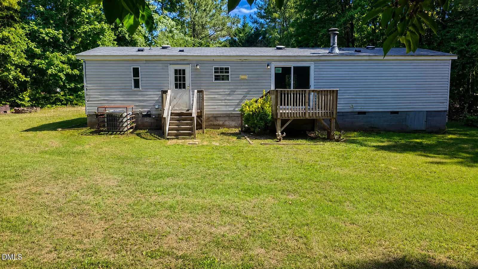 A single-story white mobile home with two small wooden porches sits on a grassy yard, surrounded by trees and greenery under a bright blue sky.