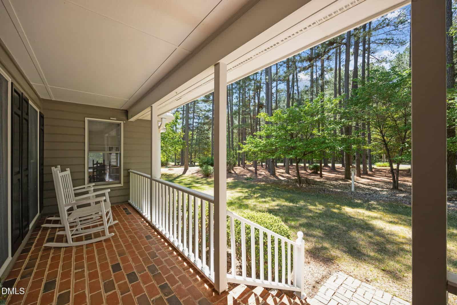 A brick front porch with white railings and two white rocking chairs overlooks a yard with green grass, trees, and a wooded area under a clear blue sky.