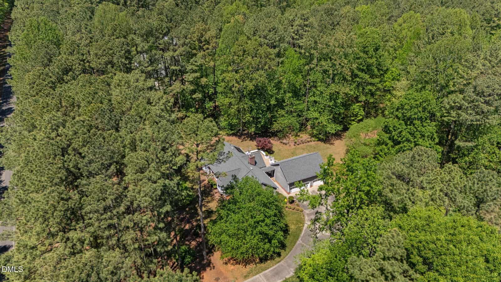 Aerial view of a house surrounded by dense trees and greenery, with a curved driveway leading to the home, located in a wooded area.