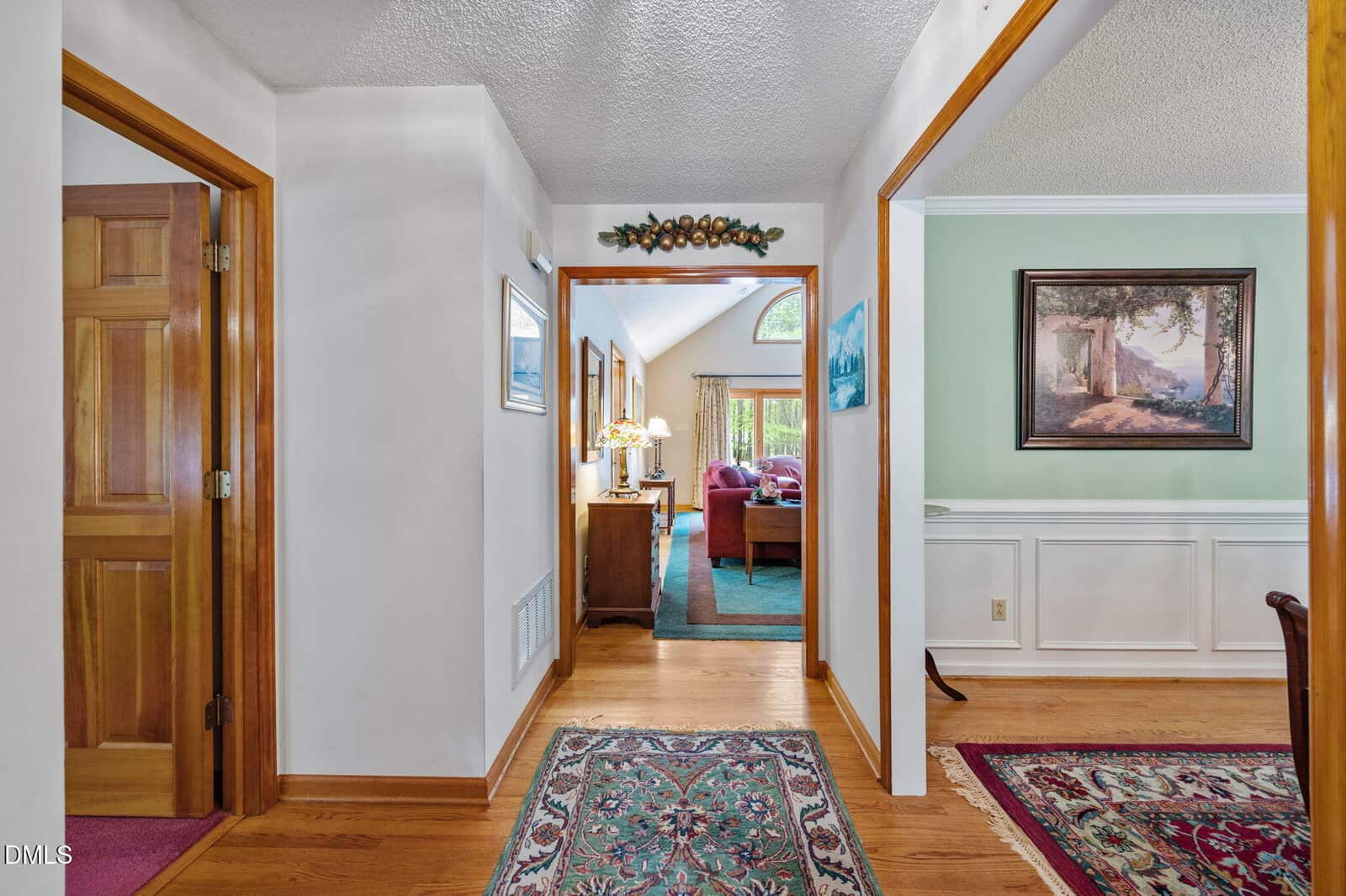 A hallway with wood floors, area rugs, white walls, and wooden doors leads to a living room with a vaulted ceiling. A decorated wall and artwork are visible, along with part of a dining room on the right.