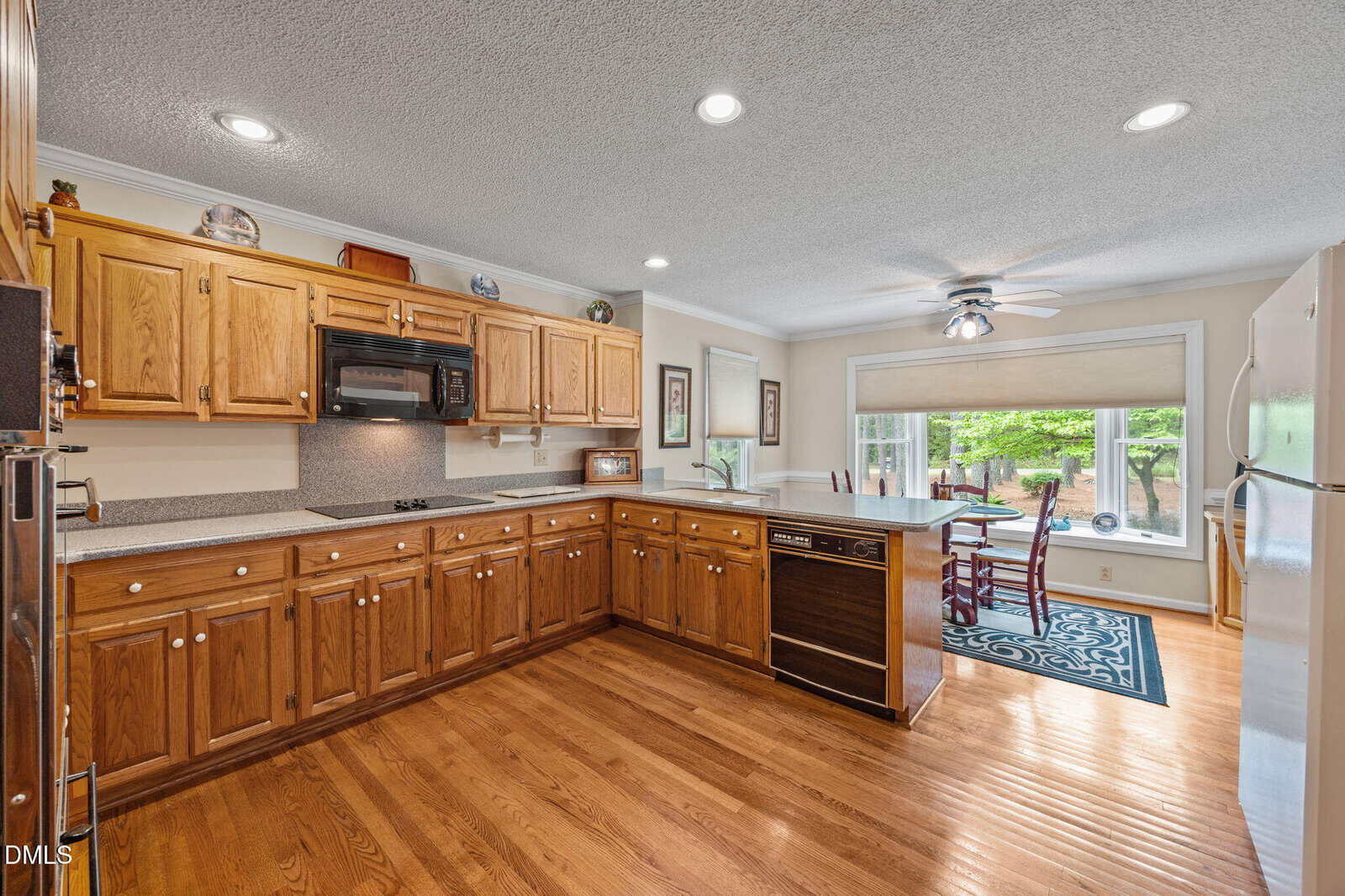 Spacious kitchen with wooden cabinets, white appliances, and hardwood floors. An island with a sink separates the kitchen from a dining area with large window overlooking a green backyard. Ceiling lights and fan are visible.