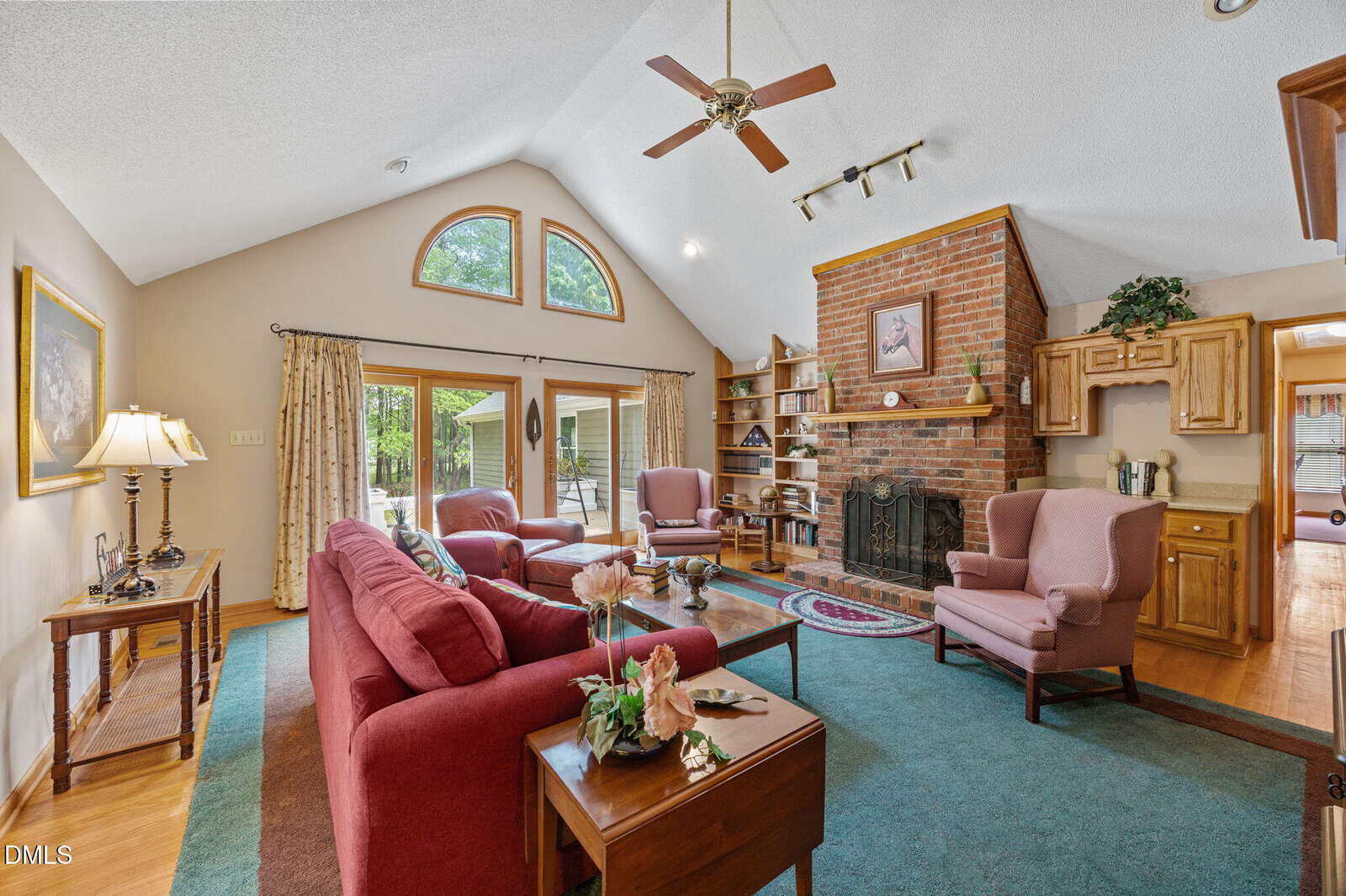 Living room with vaulted ceiling, red couches, pink armchairs, brick fireplace, wooden shelves, blue-green rug, and large windows letting in natural light. Ceiling fan and decor complete the cozy setting.