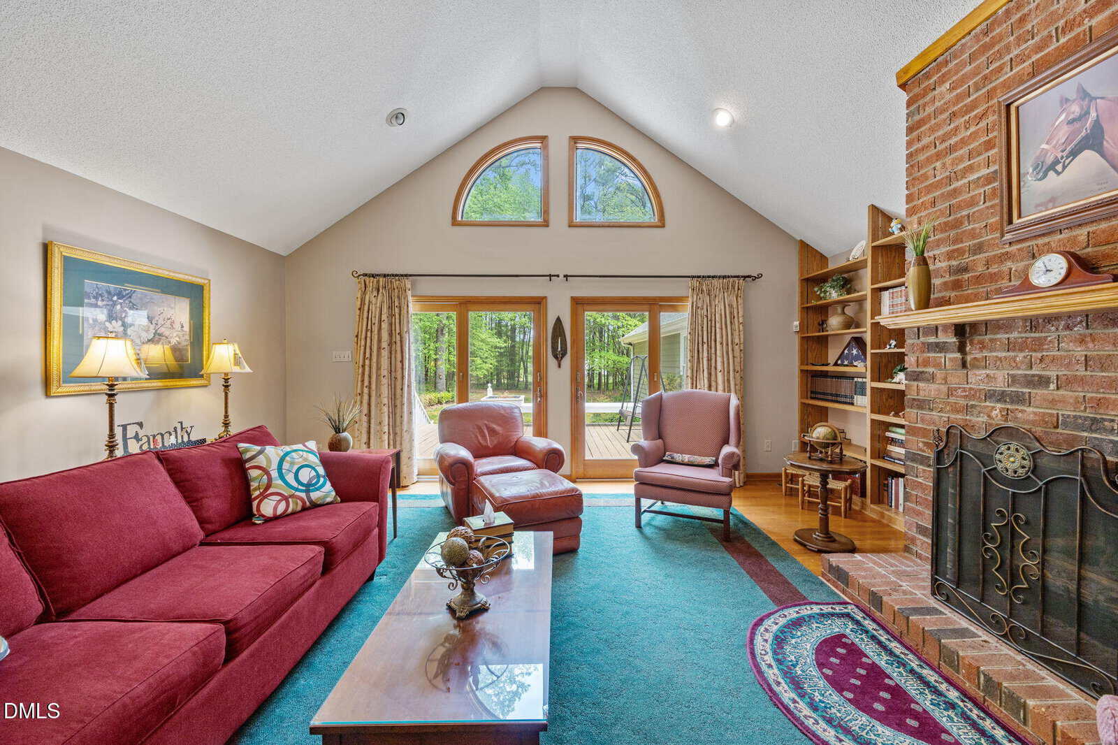 A cozy living room with red furniture, a brick fireplace, arched windows, built-in shelves, and a glass door leading outside. Decor includes framed art, lamps, and a blue-green rug.