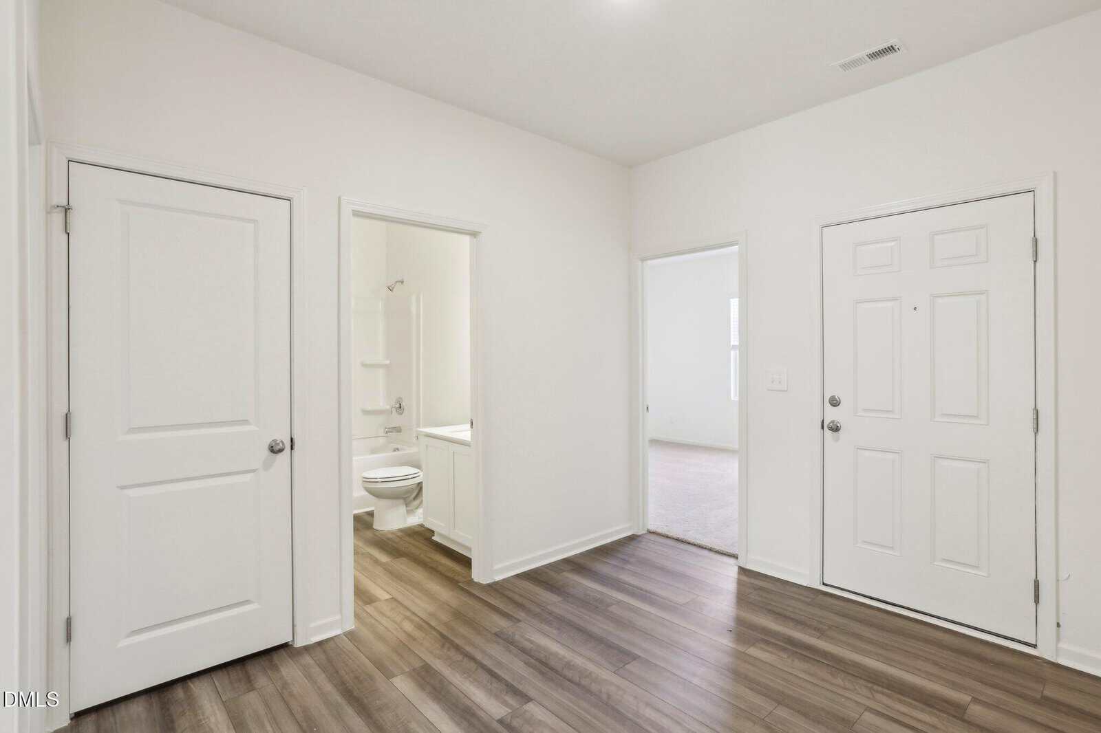 Entryway with wood flooring, white walls, and three doors: one leading to a bathroom, one to another room, and one likely a closet or utility. A white front door is on the right side of the image.