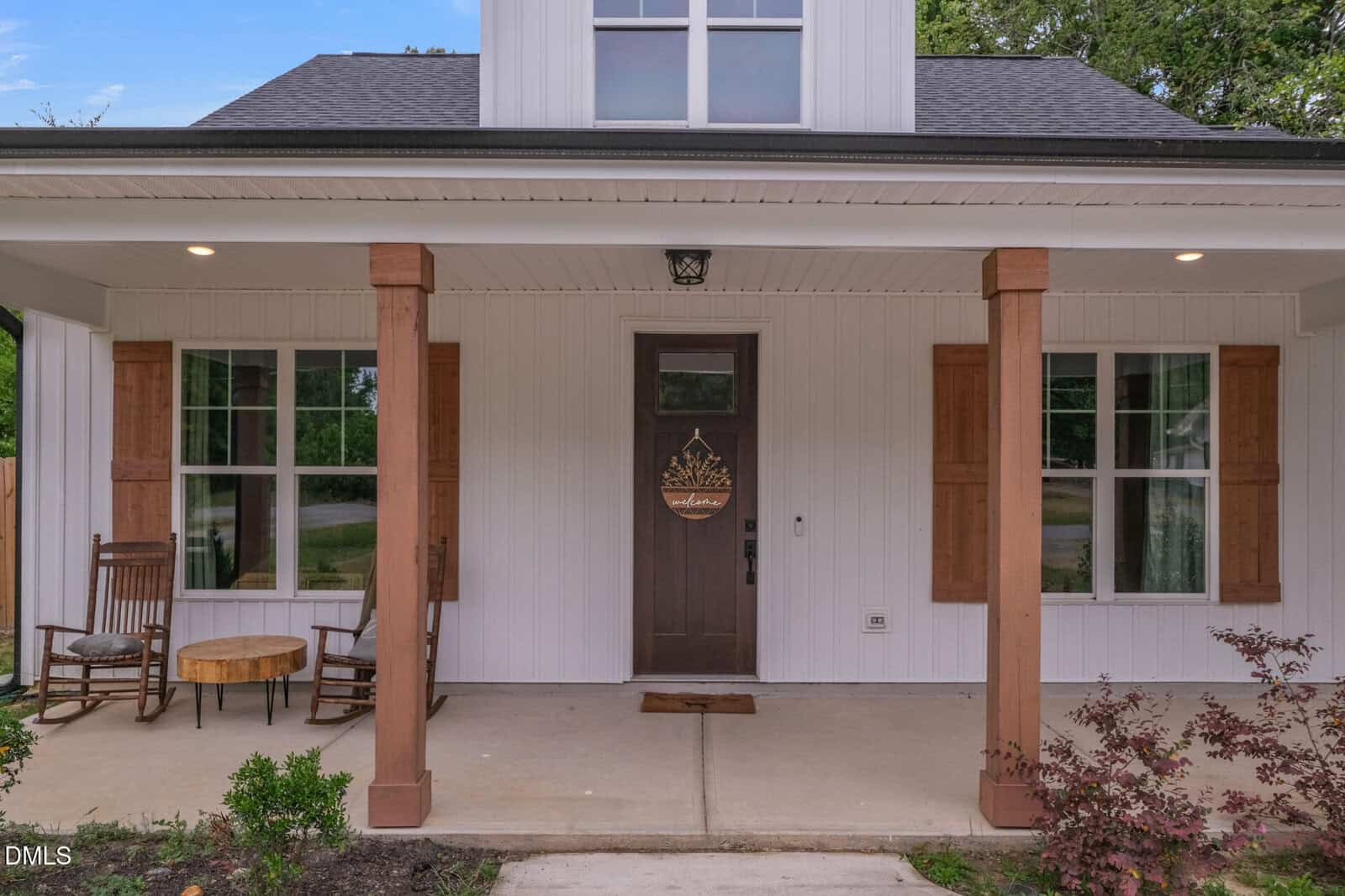 A white house with a covered front porch, wooden columns, and two windows with wooden shutters. Two chairs and a small round table sit on the left side of the porch near the front door.
