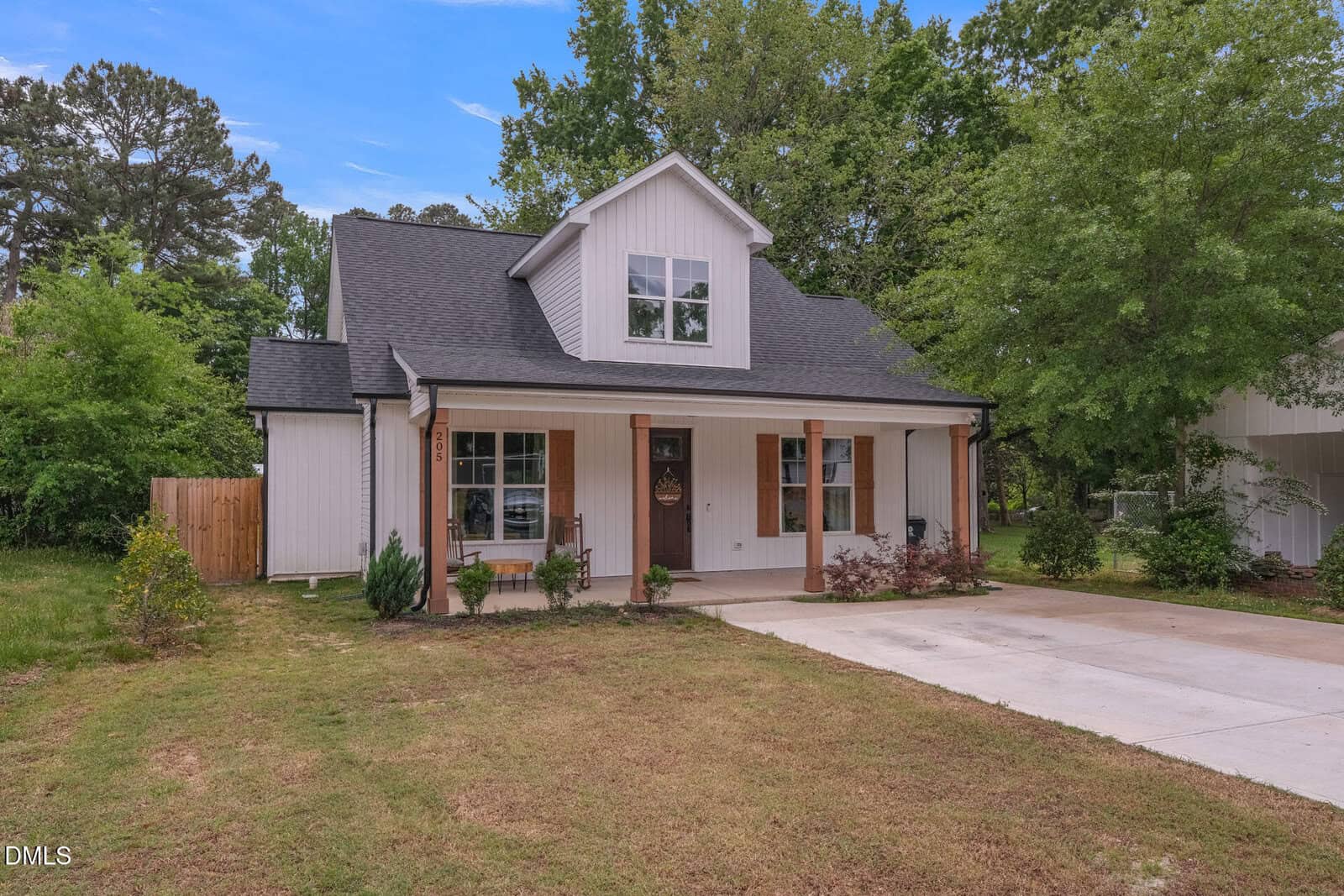 A modern two-story house with white siding, a covered front porch supported by wooden columns, and a concrete driveway. The yard has sparse grass and is bordered by trees and shrubs.