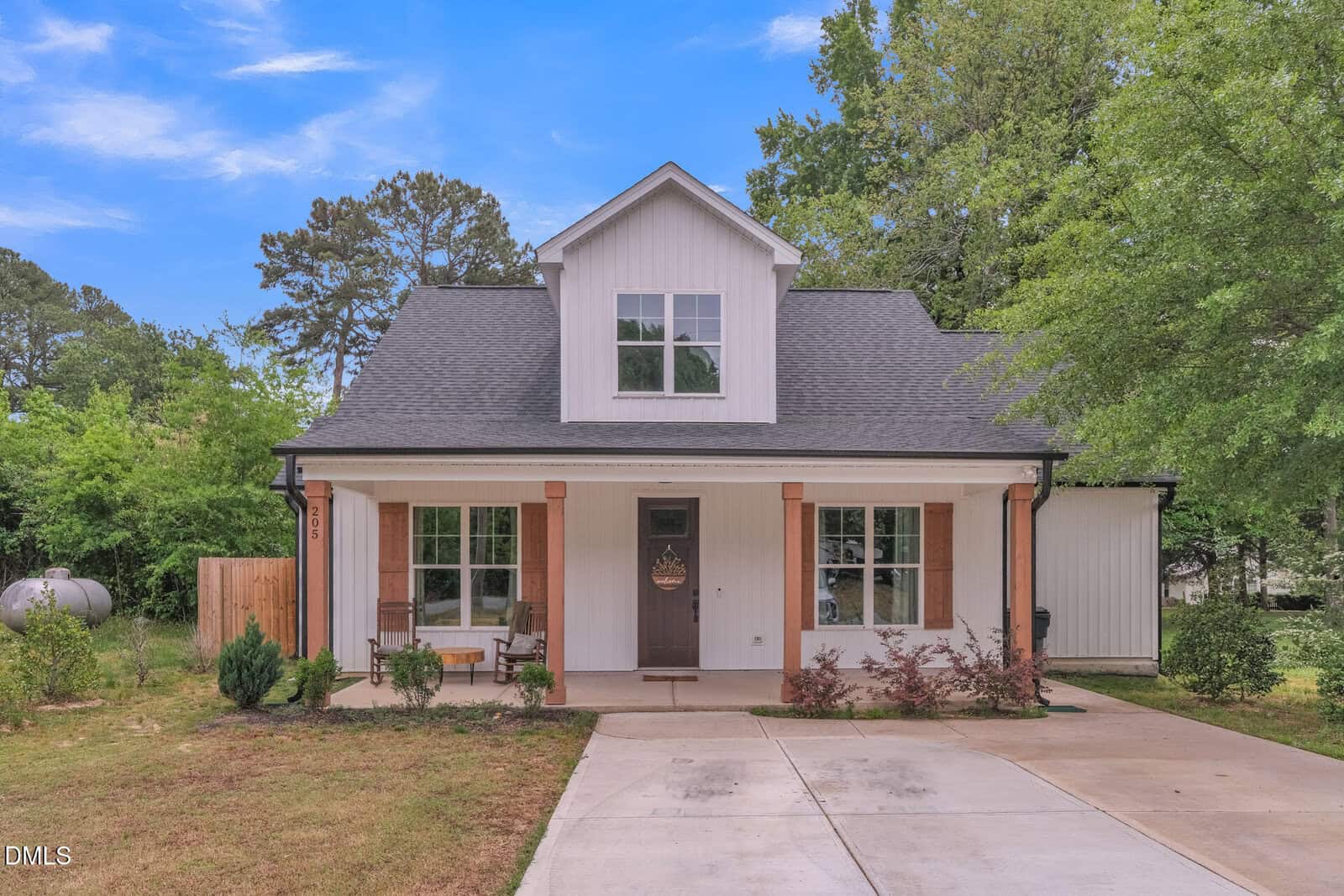 A modern, light-colored house with a dark roof, front porch, wooden columns, and a small dormer window. The driveway leads to the entrance, with a bench and some bushes in the landscaped front yard. Trees surround the home.