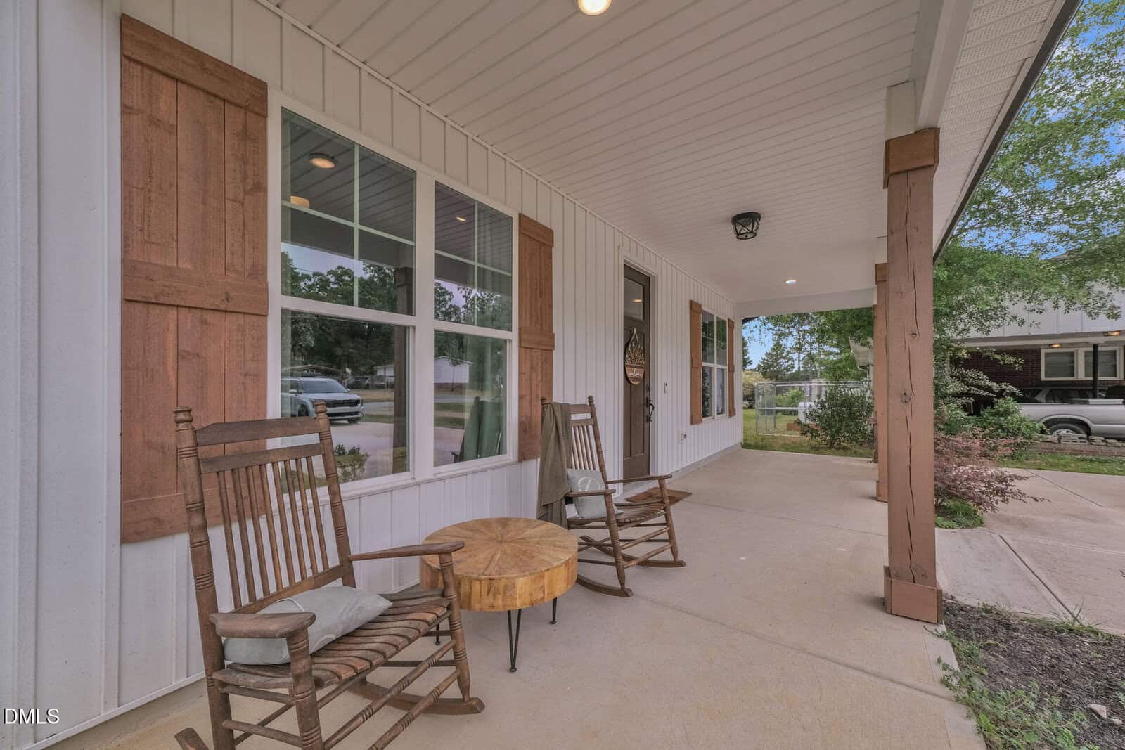 Covered front porch with two wooden rocking chairs, a small tree-stump table, decorative shutters on large windows, and greenery in the background.