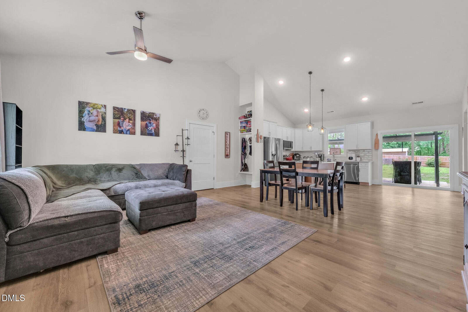 Open concept living and dining area with a gray sectional sofa, rug, dining table, modern kitchen, white walls, wood floors, and large windows with views of a green backyard.