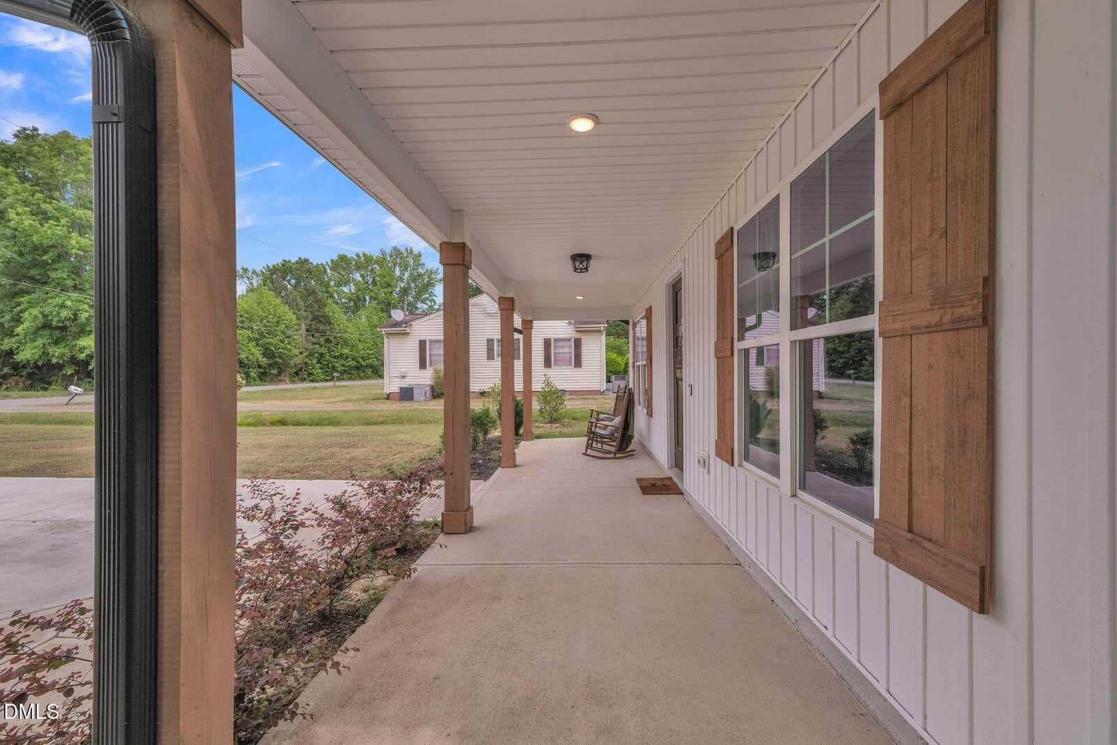 View of a covered front porch with wooden posts, rocking chairs, a doormat, exterior lights, and large windows with wood shutters overlooking a yard with trees and a driveway.
