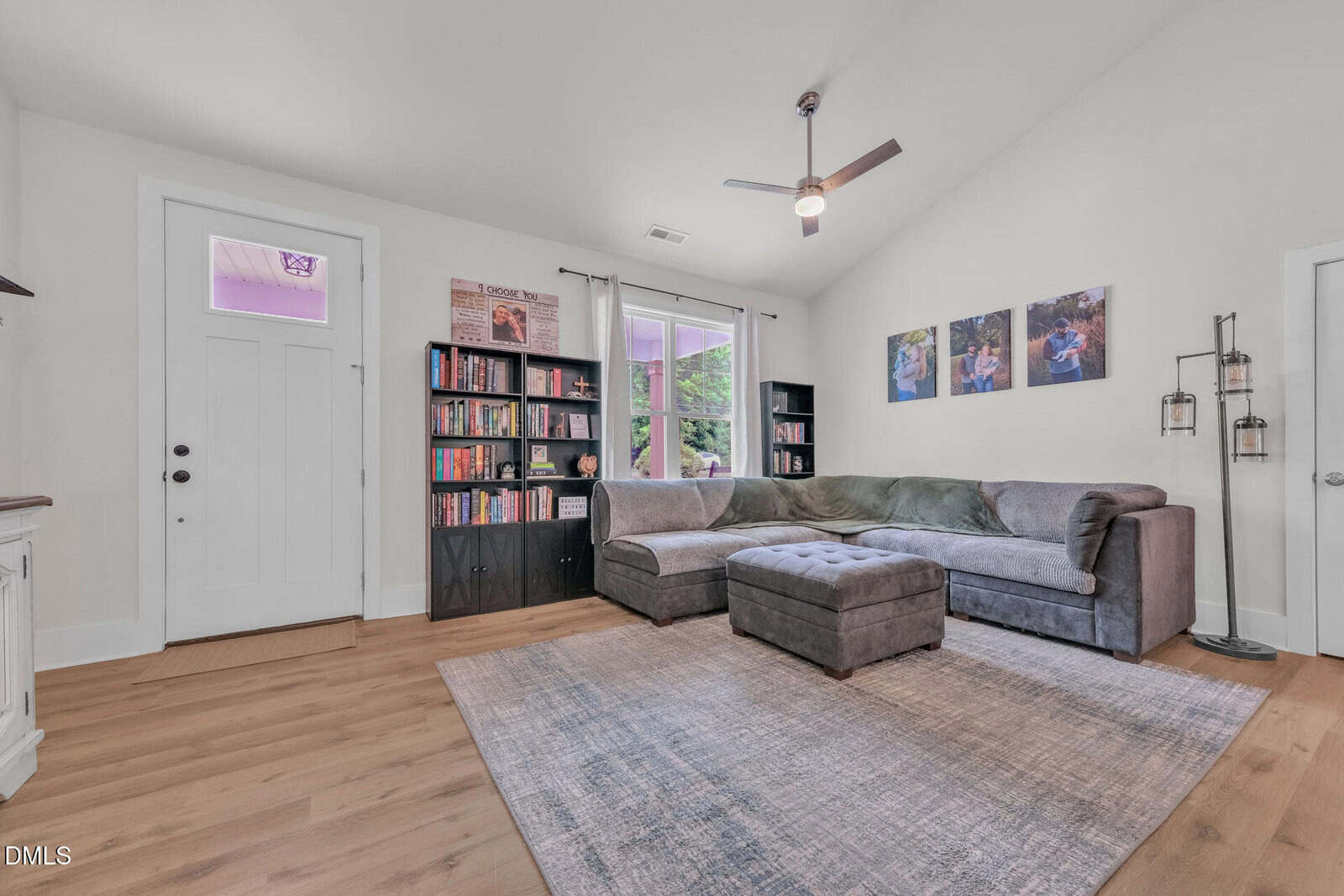A bright living room with hardwood floors, a gray sectional sofa, bookshelves, a rug, wall art, large window, ceiling fan, and a white front door.