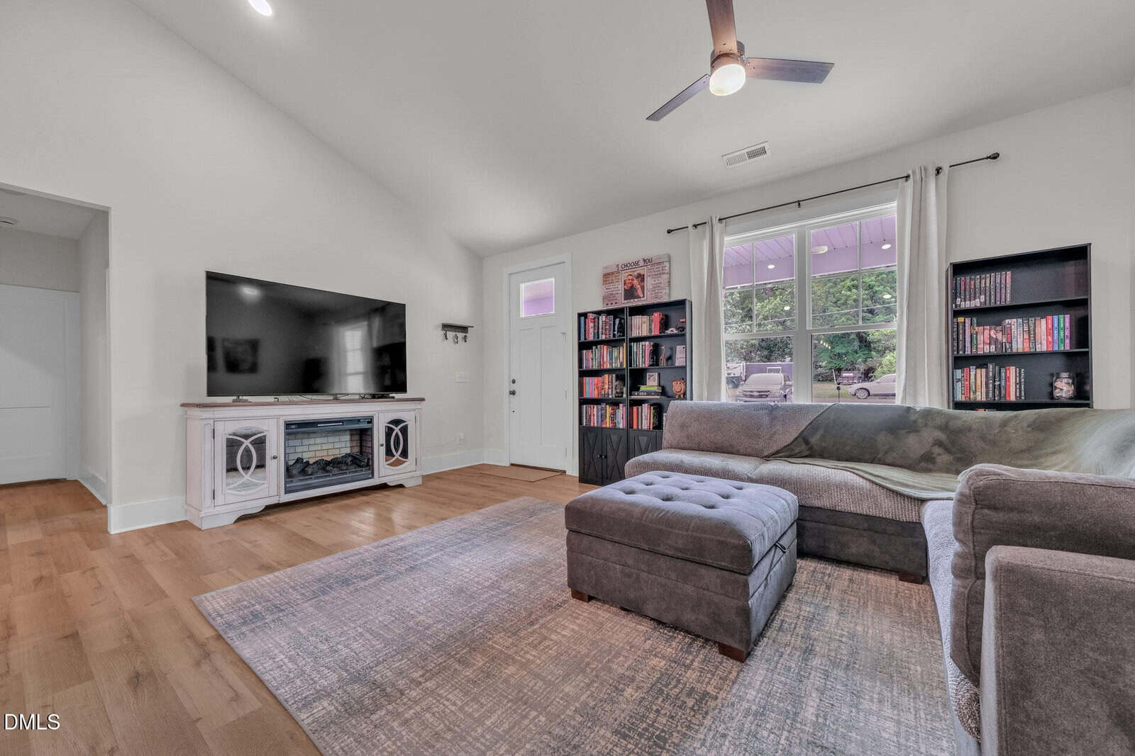 Modern living room with hardwood floors, gray sectional sofa, ottoman, large TV on a white console, bookshelves, and a window with curtains letting in natural light. Ceiling fan and area rug complete the cozy space.