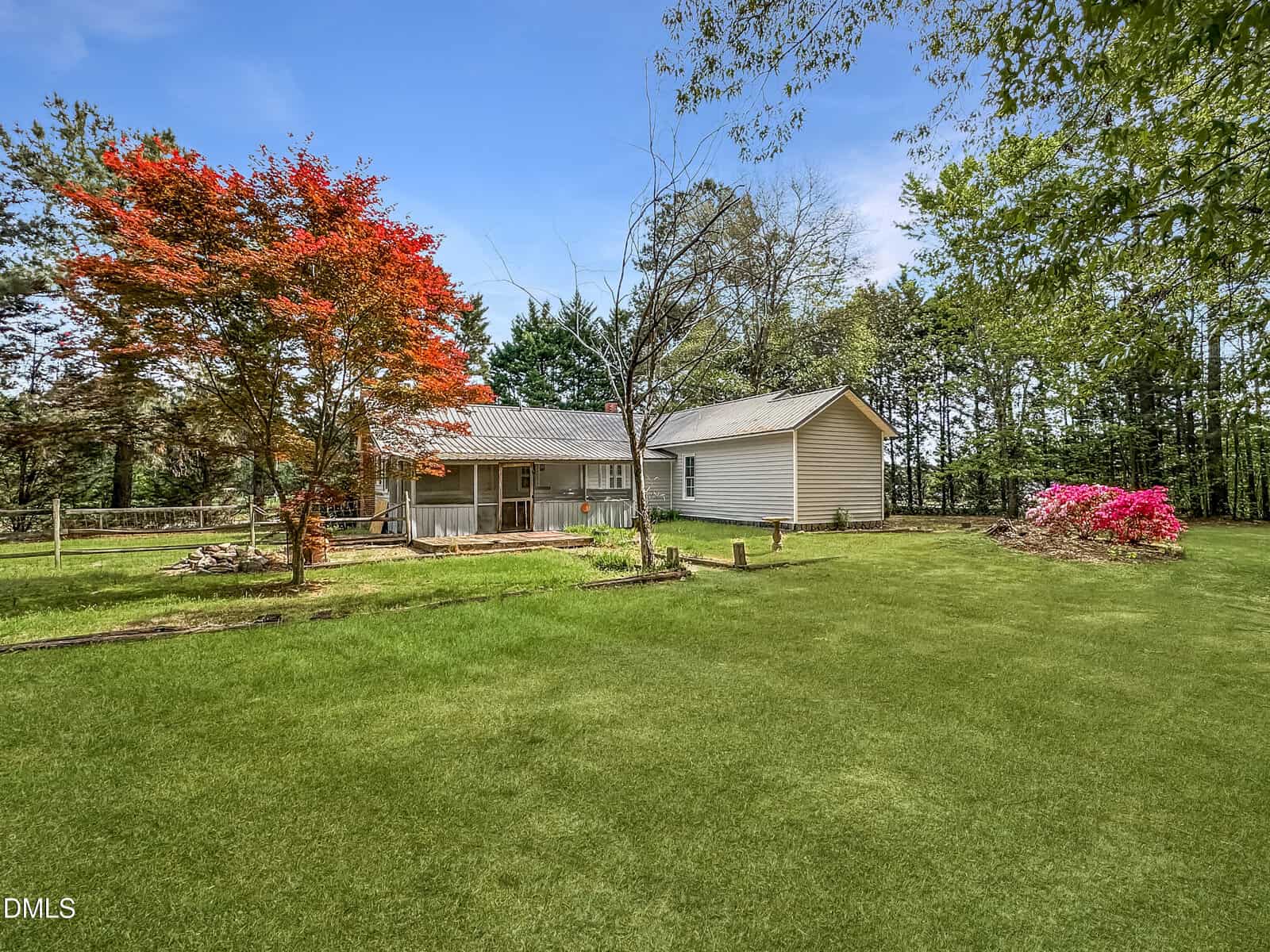 A single-story house with beige siding sits in a large, grassy yard. A tree with red leaves and a flower bed with pink blossoms are in the yard, surrounded by green trees under a blue sky.