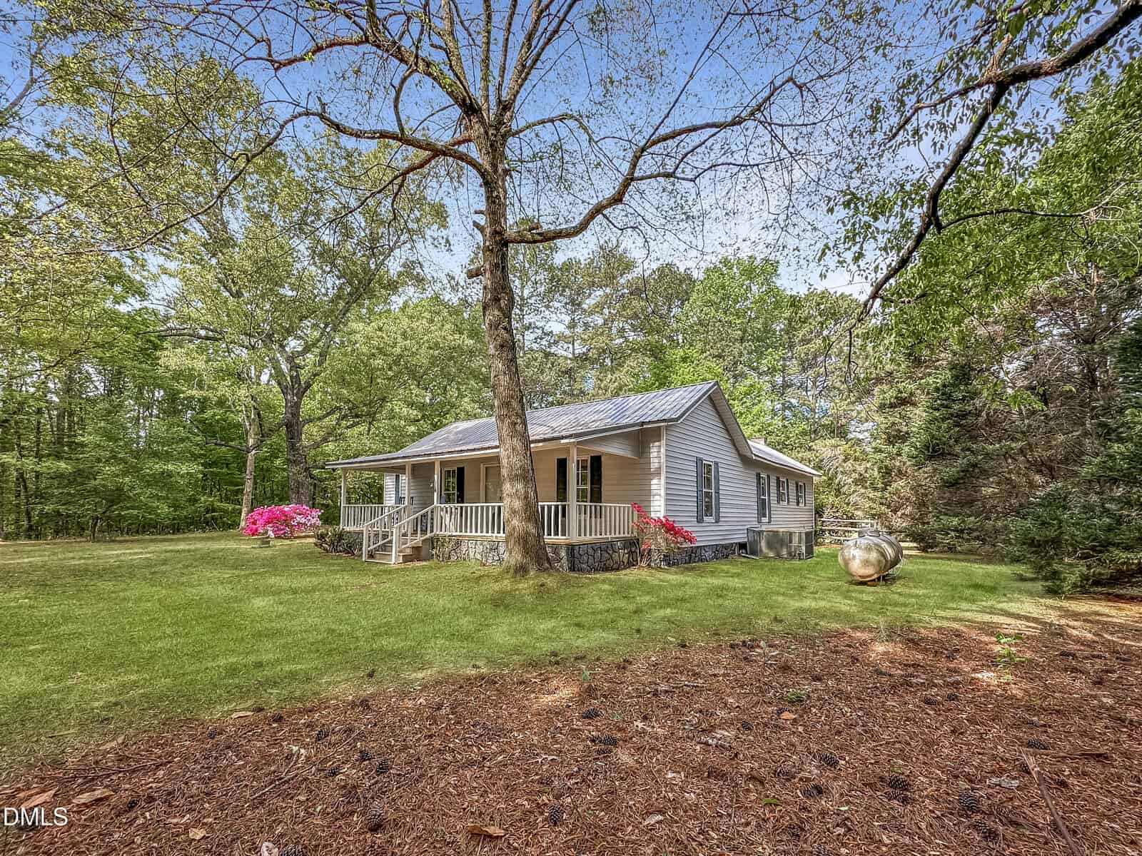 A small, light gray house with a covered front porch sits among tall trees and green grass, with pink flowers near the porch and a propane tank to the right. The scene is bright and peaceful on a sunny day.
