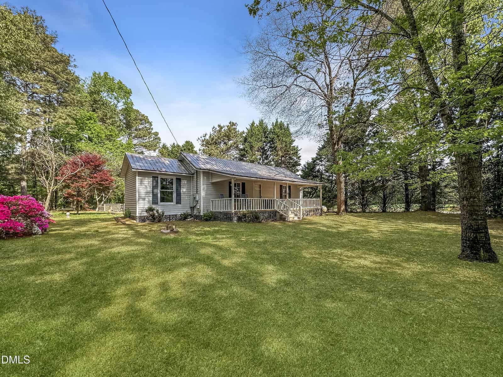 A single-story house with a porch sits on a spacious, grassy lawn surrounded by trees and blooming shrubs under a blue sky.