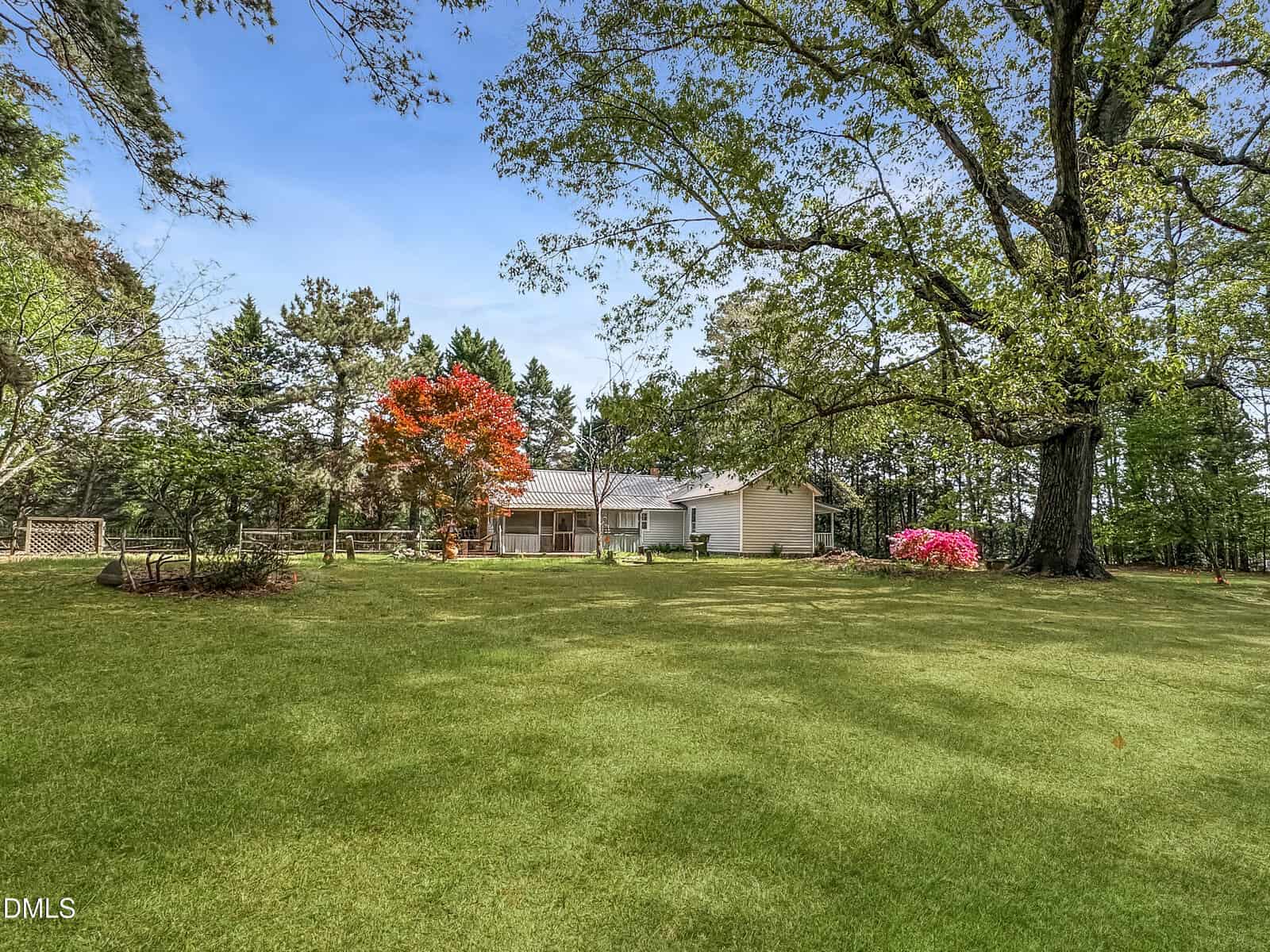 A single-story house sits among large trees and flowering shrubs on a spacious grassy lawn under a clear blue sky.