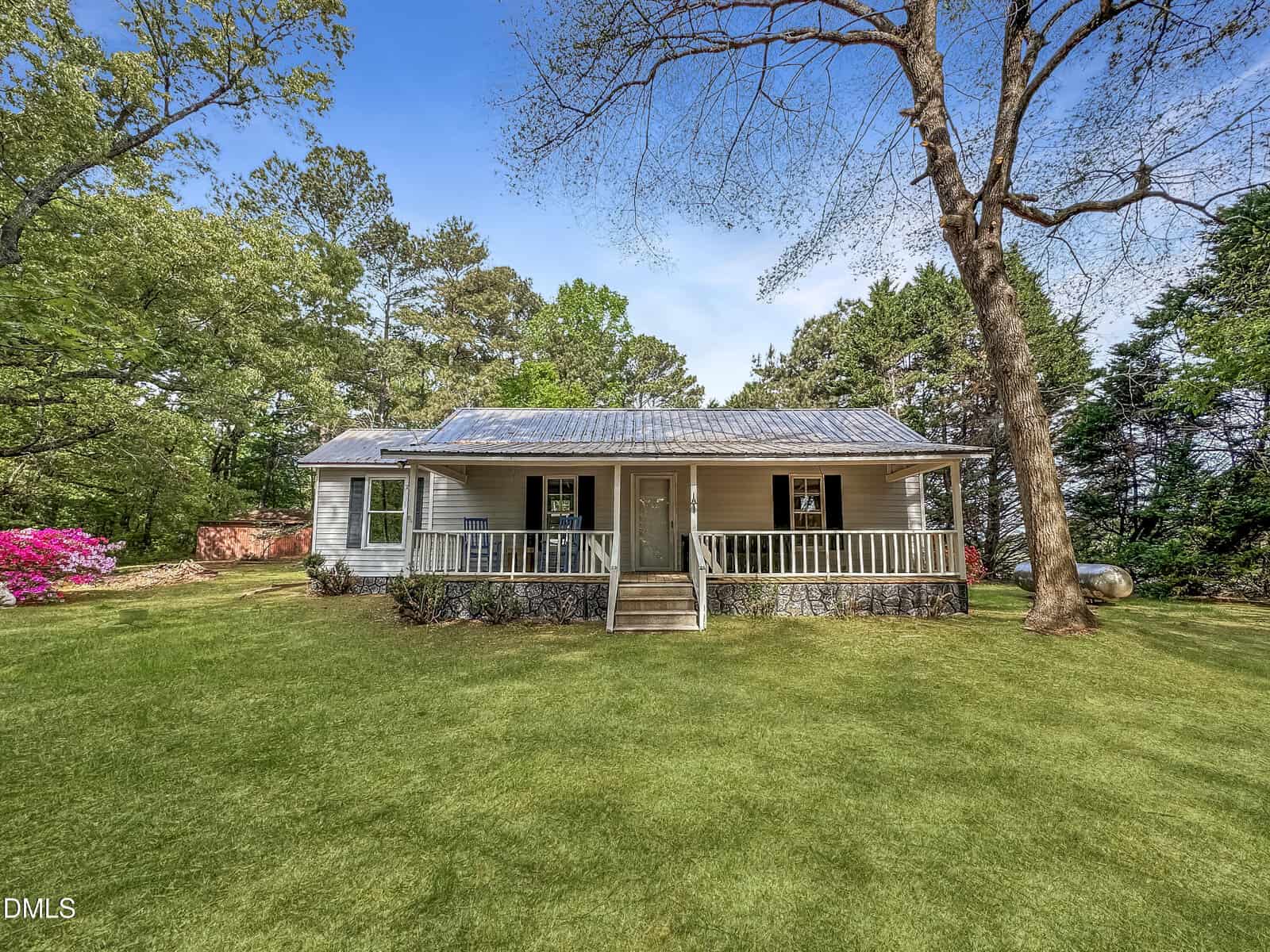 A white, single-story home with a metal roof and a wide front porch with white railings and blue rocking chairs. The house sits on a grassy lawn surrounded by trees under a bright blue sky.