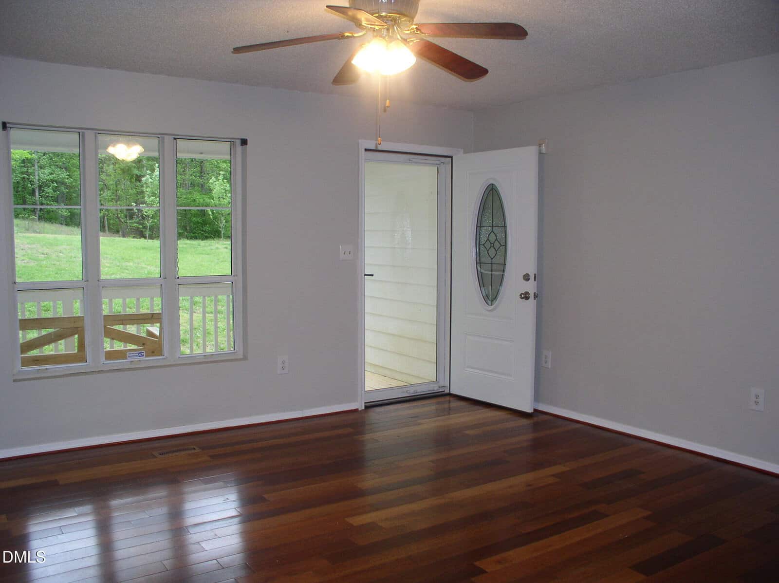 A bright, empty room with polished wood floors, light gray walls, a ceiling fan with lights, three windows, and an open white door leading to a small porch area outside.