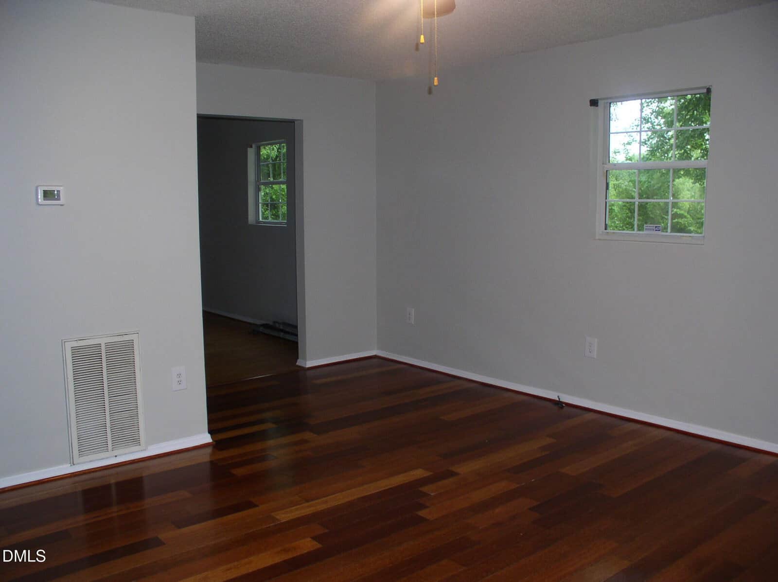 A room with smooth wooden floors, light gray walls, two small windows, a ceiling fan, and a vented panel; an open doorway leads to another room with similar flooring and a window.