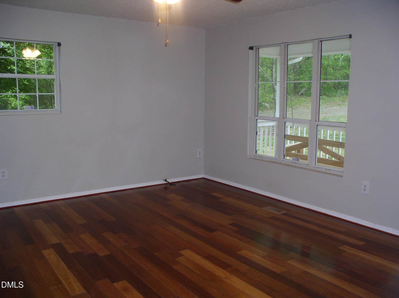Empty room with light gray walls, dark wood flooring, two windows letting in natural light, and a ceiling fan with light. A porch with a white railing is visible through the larger window.