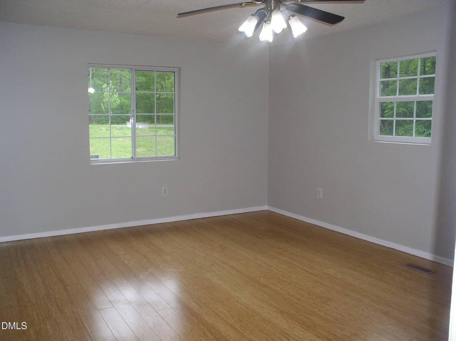 A bright, empty room with light gray walls, two windows showing green trees outside, a ceiling fan with lights, and light wood flooring.