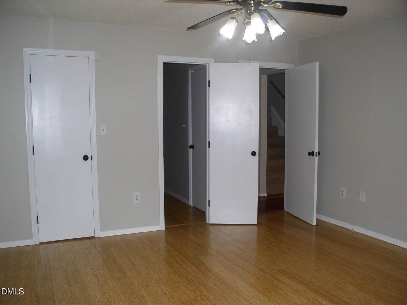 Empty room with light gray walls, wood floor, white doors, a ceiling fan with lights, and visible carpeted stairs through one doorway.