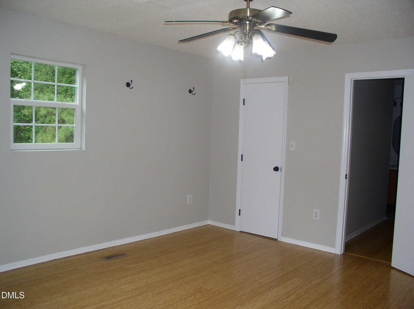 A simple, empty room with light gray walls, wood floor, white door, small window, ceiling fan with lights, and two wall hooks. Doorway leads to another room.