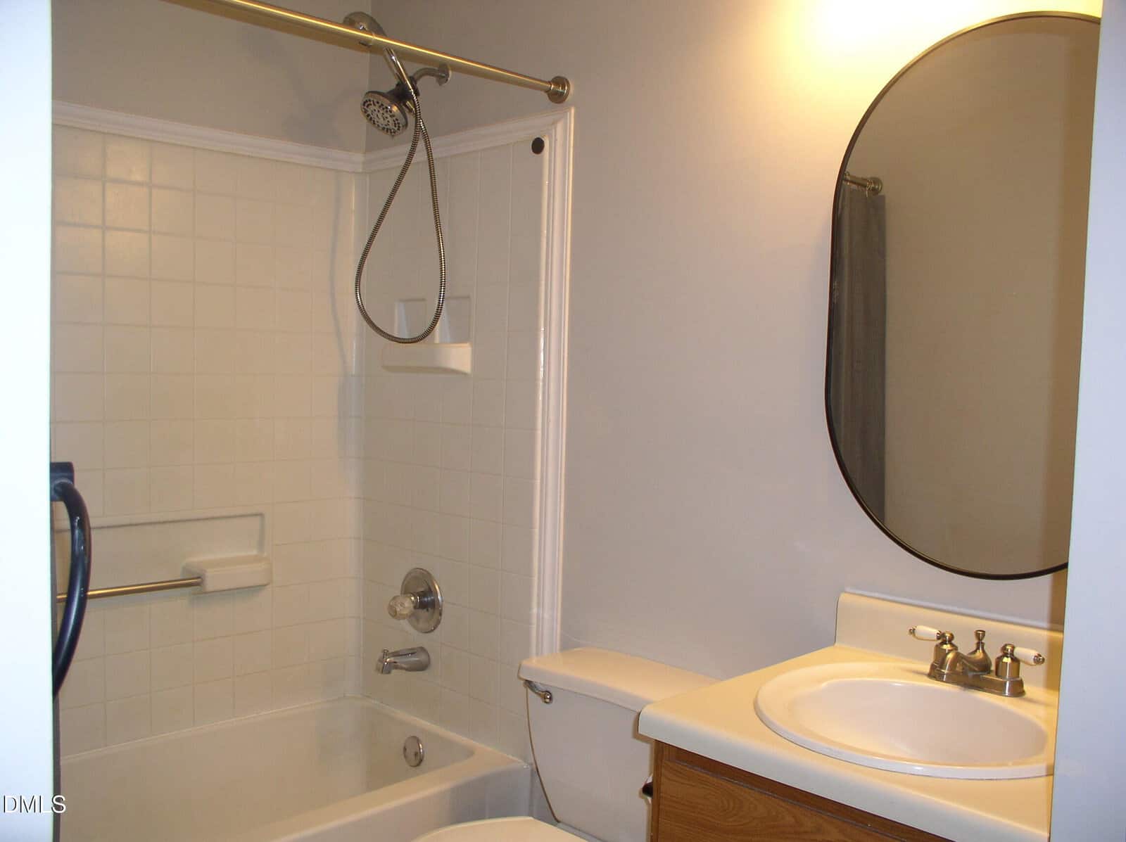 A small bathroom with a white tiled bathtub-shower combo, a showerhead, a toilet, a wooden vanity with a white sink, a mirror, and light gray walls.