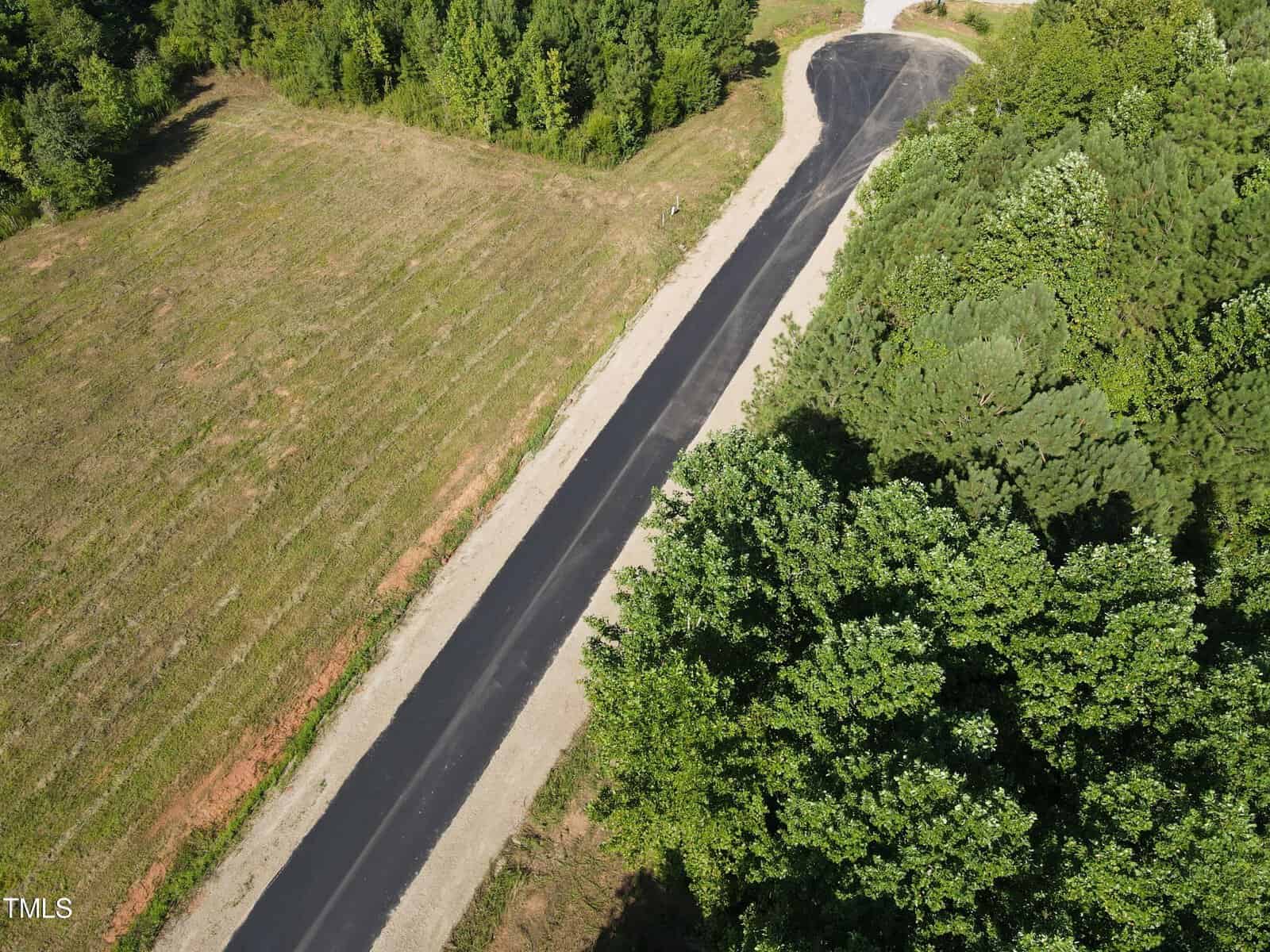 Aerial view of a newly paved road surrounded by green trees and a grassy field, leading to a small curve in the distance.