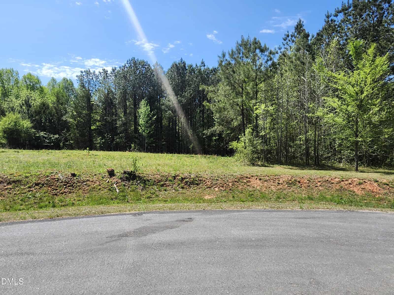 A paved road curves in front of a grassy lot bordered by tall green trees under a sunny blue sky, with a sunbeam visible across the scene.