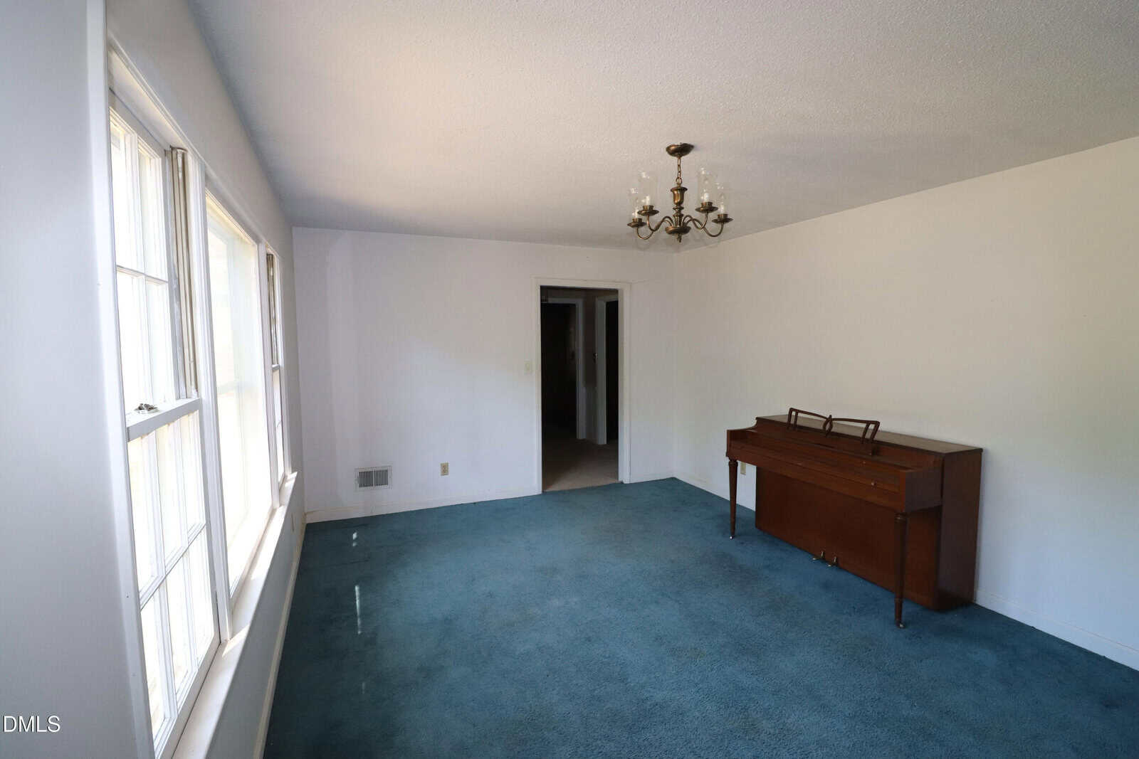 A room with blue carpet, white walls, three windows on the left, a small chandelier, and a wooden piano against the right wall. There is a doorway at the far end leading to another room.