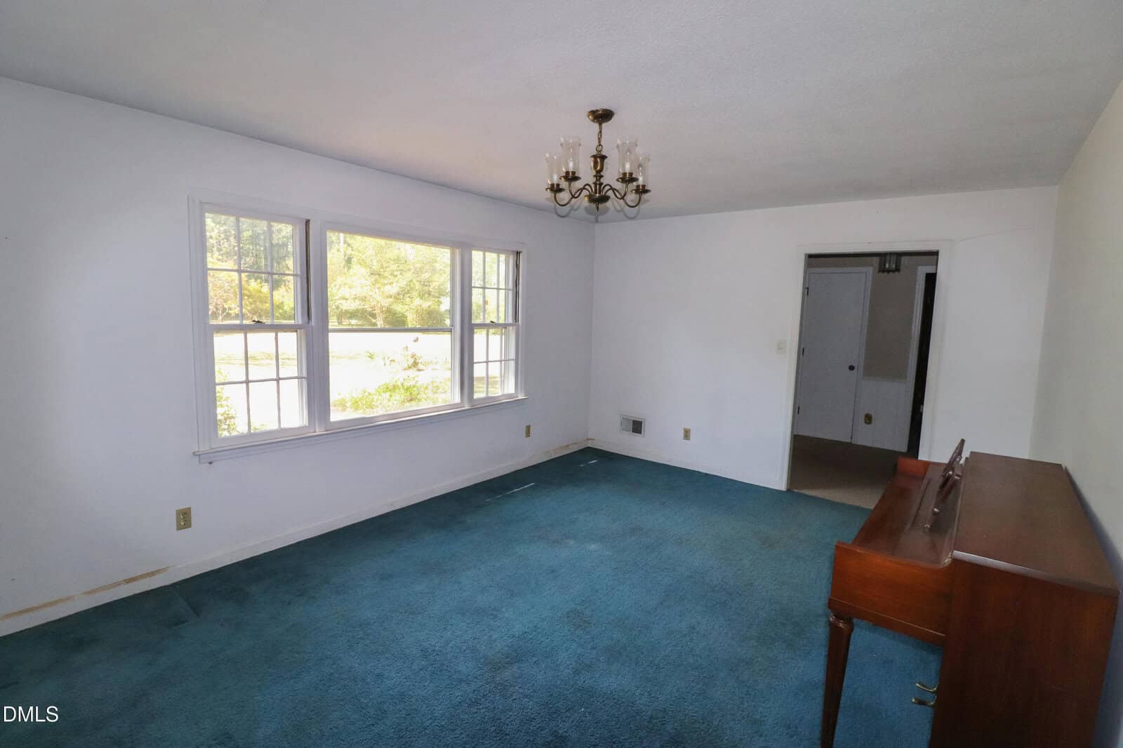 A living room with blue carpet, white walls, a chandelier, a large window letting in sunlight, and an upright piano against the right wall. A doorway leads to another room.