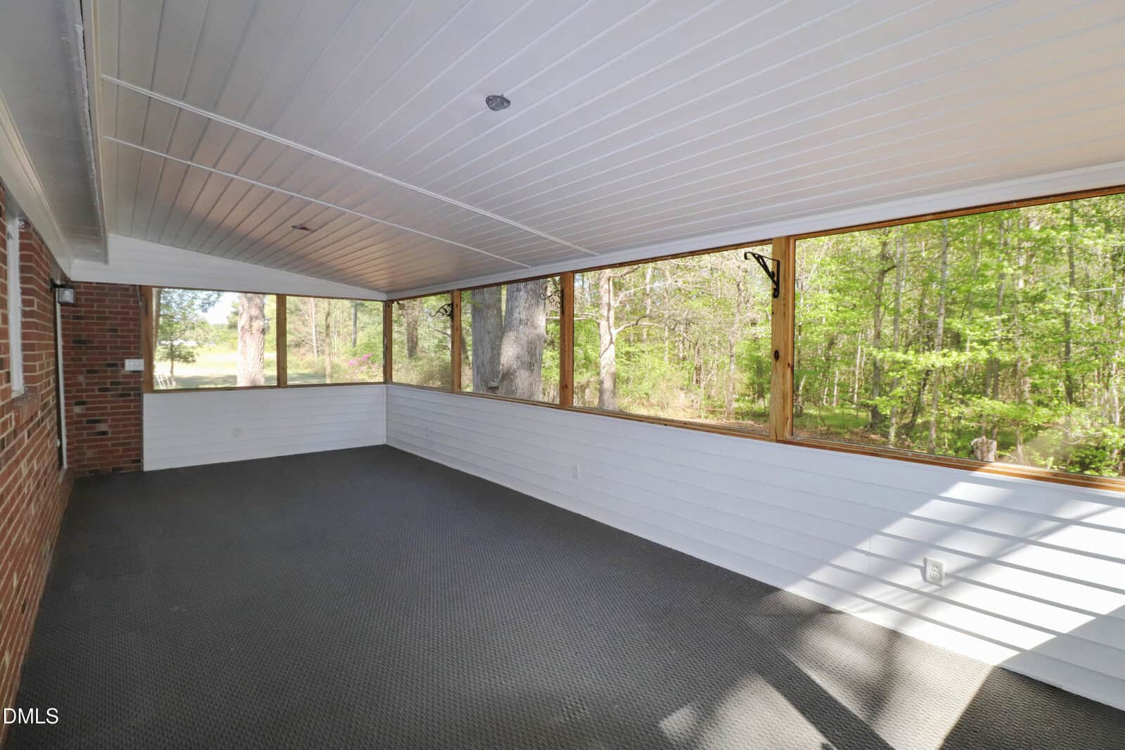 A spacious screened-in porch with a white ceiling, brick wall on one side, large screened windows, and a view of green trees outside. The floor is covered with dark carpet tiles.