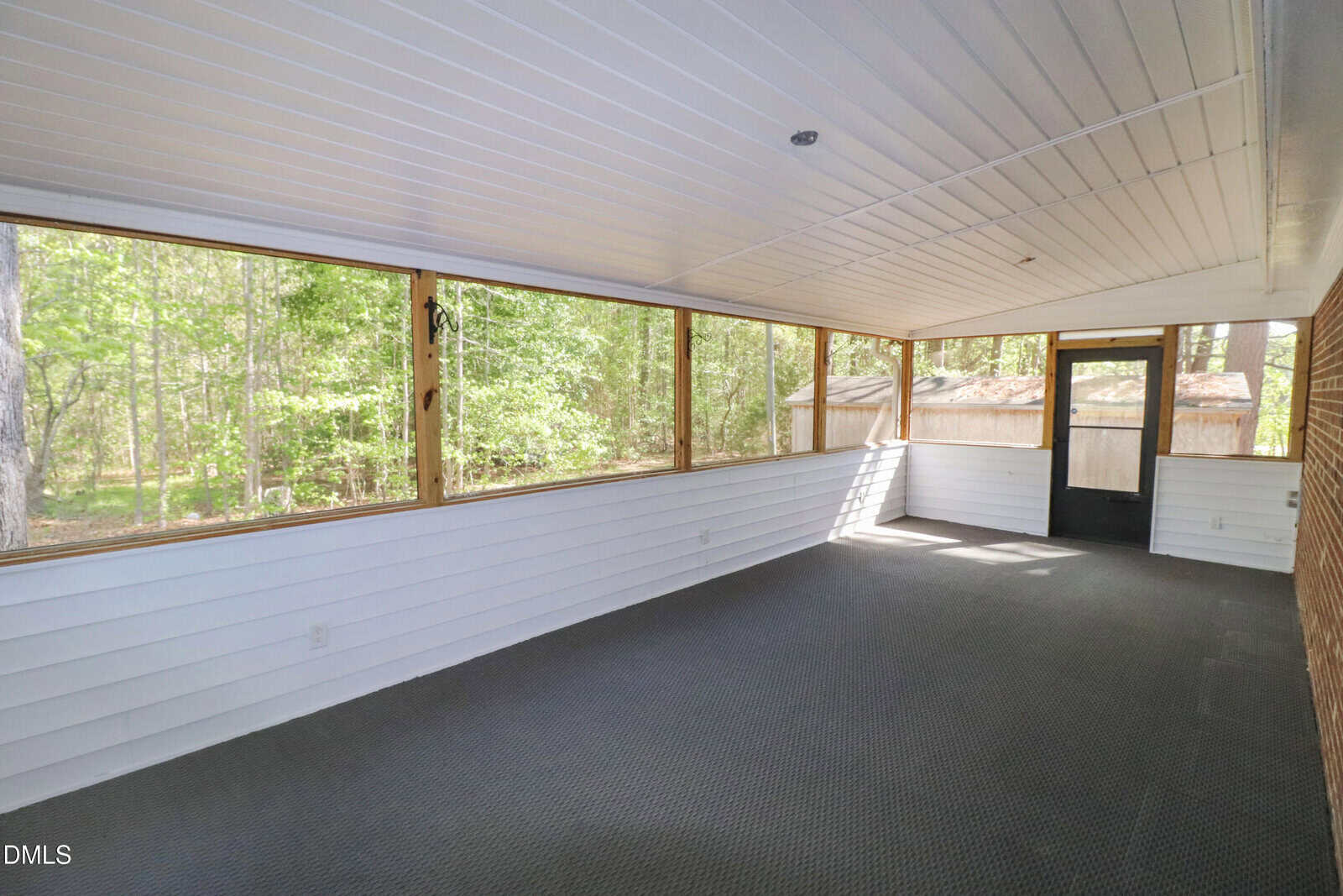 A spacious screened-in porch with white paneled walls and ceiling, dark carpeted floor, and large windows offering a view of green trees outside. A black door leads to the backyard.