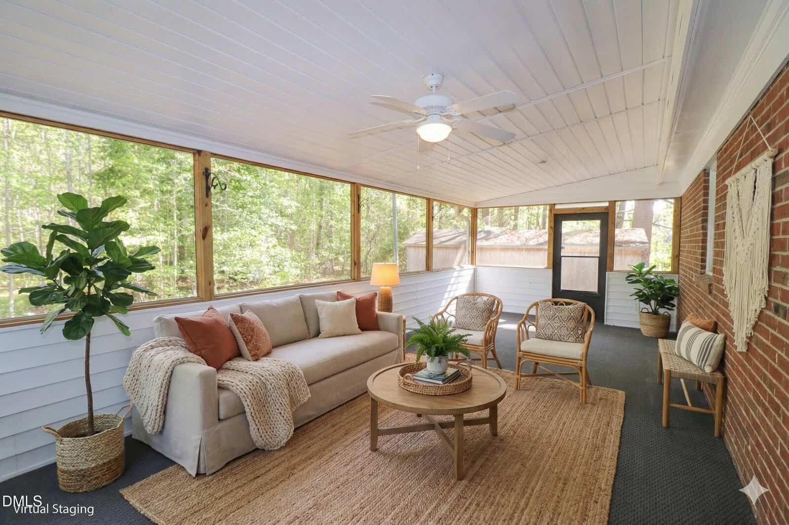 A bright sunroom with large windows, white ceiling, and brick accent wall. It features a sofa with throw pillows, two wicker chairs, a round coffee table, plants, and a woven rug. Trees are visible outside.