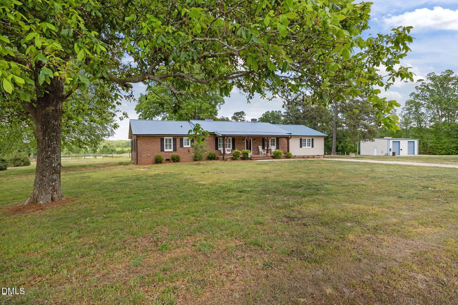 A one-story brick house with a metal roof sits on a large grassy yard with a tree in front. There is a driveway leading to a white shed or garage in the background. The sky is partly cloudy.